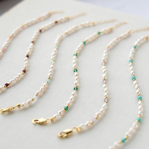 Könnte beinhalten: Eine Sammlung zarter Perlenketten mit goldenen Verschlüssen und Akzenten. Jede Halskette hat kleine, runde Perlen, die mit farbigen Edelsteinen, darunter Rot, Grün und Türkis, durchsetzt sind. Die Halsketten werden auf einer hellen Oberfläche präsentiert.