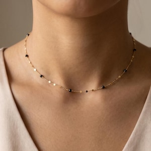 May include: A delicate gold chain necklace featuring small, evenly spaced black beads and flat gold discs. The necklace sits closely around the neck as a choker, showcasing a minimalist design with alternating metallic and dark circular accents.