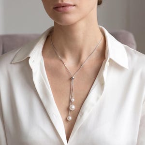 May include: A silver-toned lariat necklace featuring a delicate chain with an adjustable sliding bead and three dangling white pearls of varying sizes. The necklace is worn with a crisp, white button-down shirt, highlighting the metallic finish.