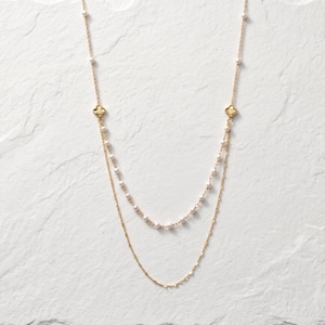 May include: A gold-toned double-strand necklace featuring a delicate chain with small white pearls and two symmetrical gold-toned clover-shaped charms. The necklace is designed with a layered appearance, combining a pearl-accented strand and a plain chain.
