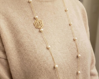 May include: A long gold chain necklace featuring evenly spaced white pearls and a central gold-toned floral charm with an openwork design. The necklace is worn over a beige knit sweater, highlighting the contrast between the metal and pearls.