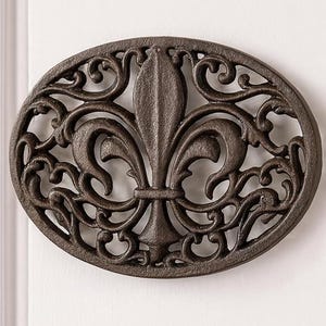 May include: Two oval, dark brown metal wall decorations with a fleur-de-lis design. The decorative pieces are mounted vertically on a white wall. The fleur-de-lis is the central design element.