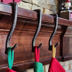 May include: Four decorative metal stocking holders hang from a dark wood mantel. Red, green, and gold Christmas stockings are suspended from the holders. A red sign with the word "Christmas" is visible above the mantel.