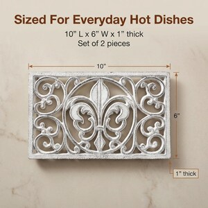 May include: White rectangular trivet with an ornate fleur-de-lis design. The trivet is 10 inches long, 6 inches wide, and 1 inch thick. The text "Sized For Everyday Hot Dishes" is at the top. Set of 2 pieces.