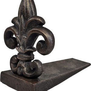 May include: Bronze-colored fleur-de-lis doorstop. The doorstop features a detailed fleur-de-lis design atop a rectangular wedge. The doorstop is made of a textured, possibly cast, material. The doorstop is designed to hold a door open.