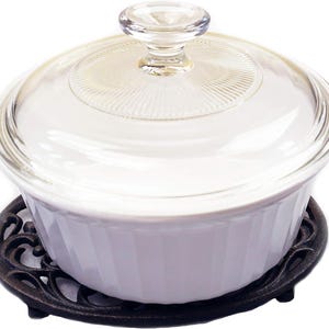 May include: A white, ribbed casserole dish with a clear glass lid and a glass knob. The dish sits on a decorative, dark brown cast iron trivet. A kitchenware item.
