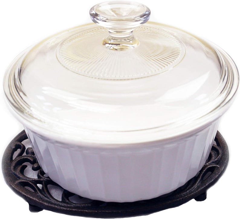 May include: A white, ribbed casserole dish with a clear glass lid and a glass knob. The dish sits on a decorative, dark brown cast iron trivet. A kitchenware item.