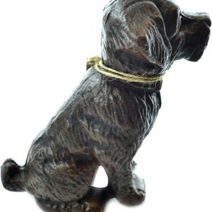 May include: A dark brown, textured cast iron dog figurine in a seated position, facing to the right. The dog features a detailed, rugged coat and is adorned with a simple, thin, light-colored twine string tied loosely around its neck.