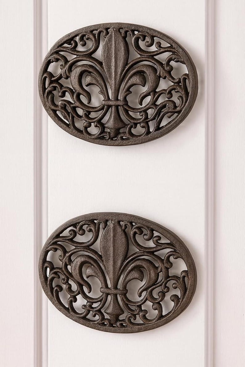 May include: Two oval, dark brown metal wall decorations with a fleur-de-lis design. The decorative pieces are mounted vertically on a white wall. The fleur-de-lis is the central design element.