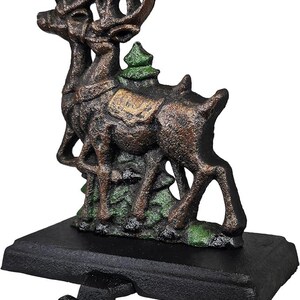 May include: A cast iron Christmas stocking holder featuring two reindeer with large antlers. The reindeer are brown with green accents and stand on a black base with a hook for hanging stockings. A small green Christmas tree is between the reindeer.