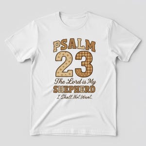 May include: White t-shirt with the text "PSALM 23" in brown, decorative lettering. The number "23" is in a patchwork design. Below the text is "The Lord is My Shepherd I Shall Not Want."