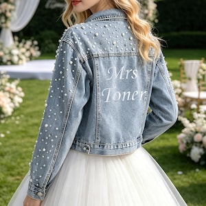 May include: A light blue denim jacket featuring white pearl embellishments scattered across the back and sleeves. The back of the jacket displays the white embroidered text "Mrs Toner". The jacket has a classic collar, long sleeves, and buttoned cuffs.