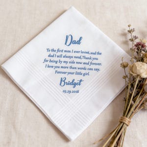 May include: A white square handkerchief with blue embroidered text that reads: Dad, To the first man I ever loved, and the dad I will always need, Thank you for being by my side now and forever. I love you more than words can say. Forever your little girl. Bridget 05.29.2016.