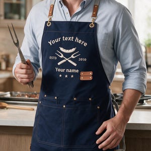 May include: A navy blue apron featuring tan leather straps with gold-tone metal hardware. The front displays embroidered text reading "Your text here", "ESTD 2010", and "Your name", along with a graphic of a sausage and two crossed forks.