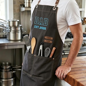 Peut inclure: Un tablier gris foncé avec des lanières en cuir marron et des boucles en métal. La poitrine porte l'inscription DAD EST.2026 en bleu clair. Le tablier possède une grande poche avant contenant une spatule, une cuillère, une pince, un fouet et un couteau.