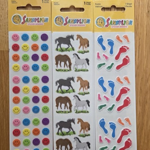 May include: Three packs of Sandylion sticker designs. One pack features colourful smiley faces, another has horse illustrations, and the third displays footprints. Each pack has the text "Fuzzy Velours" and "Sticker Designs".
