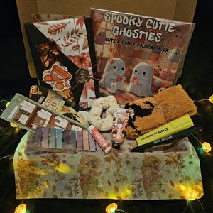 May include: A gift box containing a "Spooky Cutie Ghosties" colouring book, orange socks, a scrunchie, and lotion. The box is decorated with autumn patterns and fairy lights. The box is filled with various items.