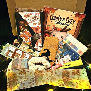 May include: An open cardboard box filled with autumn-themed items. Includes a colouring book, socks with a witch design, a bookmark, a scrunchie, and stationery. The box is decorated with autumn leaves.
