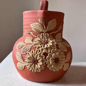 May include: A terracotta-coloured ceramic vase with a handle and floral relief details. The vase features three large flowers with tan petals and red centres, along with leaves and buds. The vase has a textured surface.