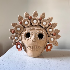 May include: A tan ceramic skull sculpture with floral and leaf accents. The skull has large eye sockets, a smiling mouth, and is adorned with flowers in shades of tan, white, and orange. The sculpture is a decorative piece.