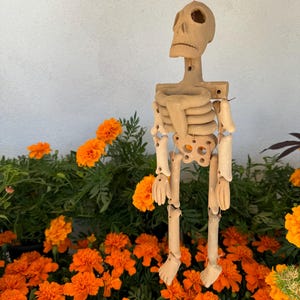 May include: A hanging skeleton figurine crafted from light tan clay, featuring articulated limbs connected by thin string. The figure has a hollow skull, a rib cage, and a pelvic bone structure, suspended by a single string from the top of the head.