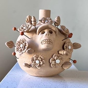 May include: A round, light brown terracotta vessel featuring a central sculpted skull wearing a cap. The surface is decorated with applied clay flowers and leaves, accented by small white circular centers and thin metal pins with orange tips.