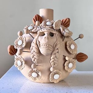 May include: A round, light tan clay vessel featuring a central sculpted skull with braided hair details. The surface is adorned with multiple small, white-centered clay flowers and dark terracotta-colored leaf accents attached to thin metal pins.