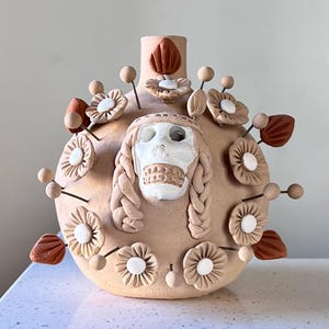 May include: A round, light brown clay vase featuring a central white skull with braided hair details. The surface is decorated with numerous small, light brown and dark terracotta floral accents and thin stems, creating a textured, layered appearance.