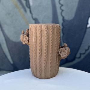 May include: A tan ceramic vase shaped like a cactus, with a textured surface and vertical ridges. It has flower and leaf embellishments. The vase is on a white surface.