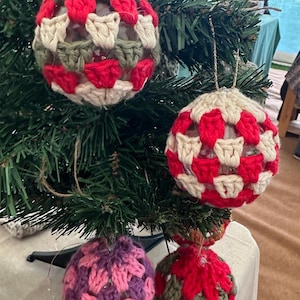 Può includere: Una collezione di decorazioni natalizie fatte a mano. Le decorazioni sono all'uncinetto in vari colori, tra cui rosso, bianco, verde e viola. Le decorazioni sono sferiche e hanno un aspetto strutturato. Sono appese a un albero di Natale verde.