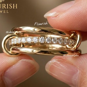 Pode incluir: Um anel de ouro com três bandas entrelaçadas, sendo que a banda central está incrustada com uma fila de pequenos diamantes redondos e transparentes. O texto FLOURISH JEWEL é visível no canto superior esquerdo, com a palavra Flourish acima.