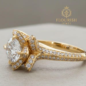 Könnte beinhalten: Ein goldener Verlobungsring mit einem großen, runden, klaren Edelstein in der Mitte. Der Ring hat kleinere Edelsteine, die entlang des Bandes und um den Mittelstein gesetzt sind. Der Ring hat den Text "Flourish Jewel".