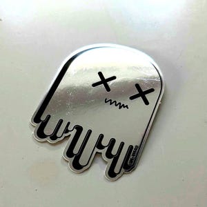 May include: A silver sticker in the shape of a ghost with a black outline. The ghost has black cross-shaped eyes and a zig-zag mouth. The bottom of the ghost has a dripping design.