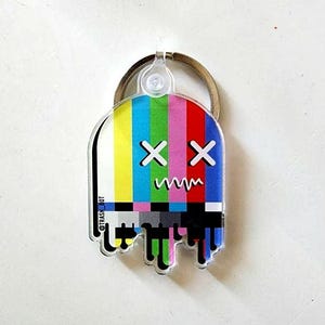 May include: A clear plastic keychain shaped like a ghost with a TV test pattern design. The ghost has two X's for eyes and a wavy line for a mouth. The text "@TRASH_BOT" is printed on the side of the keychain.