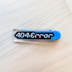 May include: A blue and clear rectangular pin with the text "404:Error" in a pixelated font. The pin has a rounded end and a black border around the text. The pin is on a light-colored surface.
