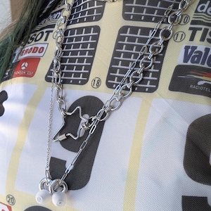 May include: A silver-toned metal chain necklace featuring a star-shaped carabiner clasp and two small white earbud attachments. The necklace is worn over a cream, yellow, and black short-sleeved shirt with various printed logos including Michelin and Elf.