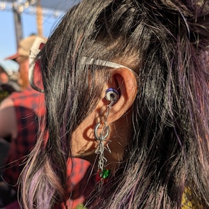May include: A person with long, dark purple and black hair wearing a silver chain earring with a star charm and a red and green charm. The earring is attached to a small, round, purple and silver ear cuff.