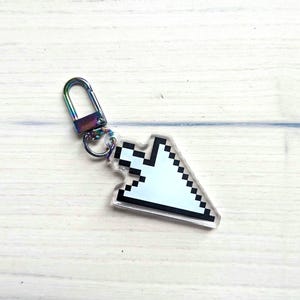 Può includere: Un portachiavi a forma di cursore del mouse pixelato, raffigurante una freccia bianca con un bordo nero. Il ciondolo è fissato a un moschettone metallico dai riflessi arcobaleno, che crea un contrasto vivace con il design digitale.