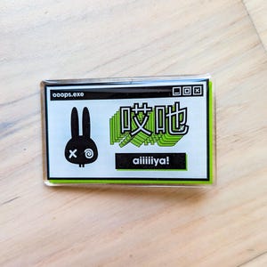 Op de afbeelding: Een rechthoekige sticker met een zwarte konijnengrafiek en de tekst "ooops.exe". De sticker bevat ook Japanse karakters en het woord "aiiiliya!" in een vetgedrukte letter, met een groen-wit kleurenschema.