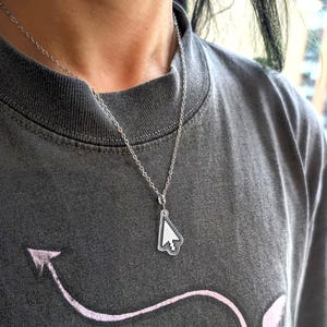May include: A silver-toned metal chain necklace featuring a small, clear acrylic pendant shaped like a computer mouse cursor. The pendant displays a white, pixelated arrow icon with a black outline, suspended from a simple, delicate link chain.
