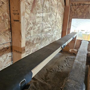 Könnte beinhalten: Eine Holzkonstruktion mit einem langen, dunkel gefärbten Streifen. Die Konstruktion besteht aus hell gefärbtem Holz und ist an einer Wand aus Holzverbundmaterial angebracht. Der dunkle Streifen hat eine Metallschlaufe und einen Riemen.