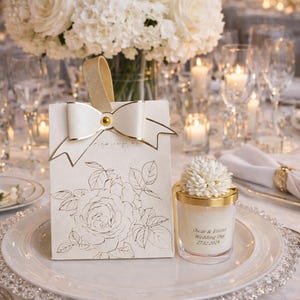 May include: A wedding table setting featuring a white floral arrangement, candles, and place settings. A gift bag with a rose design and a candle with a floral topper are on a plate. The candle has text that reads "Oscar & Emma Wedding Day 27.12.2025".