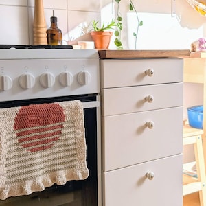 May include: A kitchen scene with a white oven, a light-colored cabinet with four drawers, and a wooden shelf. A crocheted towel with a red heart design hangs on the oven door. A wooden pepper grinder and a glass bottle are on the counter.