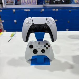 May include: A blue stand displaying a white PlayStation 5 controller above a white Xbox controller. The PlayStation controller has black detailing. The Xbox controller has coloured buttons. The controllers are for gaming.