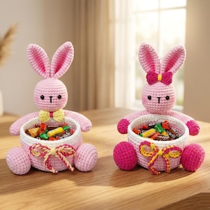 May include: Two pink crochet bunny baskets filled with candy sit on a wooden table. The bunnies have long ears, black eyes, and hold the baskets with their arms. One bunny has a yellow bow tie, and the other has a pink bow.