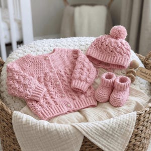 Op de afbeelding: Een roze gehaakte baby set, bestaande uit een vest, een muts met pompon en babyslofjes, gepresenteerd in een rieten mand. De set is gemaakt van zacht garen en heeft een textuur steekpatroon. Een houten speelgoed en een label met "Crochet For Life - Handmade with Love" staan ook in de mand.