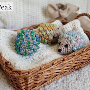 May include: A wicker basket contains three crocheted hedgehogs. Each hedgehog is decorated with colourful beads, one in green and blue, another in yellow and green, and the third in blue and pink. The basket rests on a white crocheted blanket.