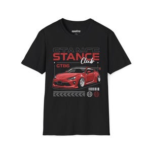 Pode incluir: T-shirt preto com um gráfico de carro esportivo vermelho e o texto "STANCE Club GT86". O design inclui uma ilustração estilizada de carro com spoiler e detalhes de corrida. A camisa tem gola redonda e mangas curtas.