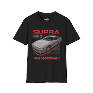 以下が含まれることがあります： グレーのSupra MK3車のグラフィックと赤い文字が特徴の黒いTシャツ。「SUPRA MK3」と「JDM LEGENDARY」の文字。クルーネックと半袖のデザインです。