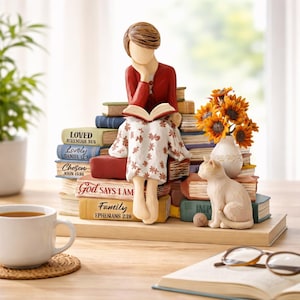 May include: A figurine of a person reading, seated on a pile of books. The figurine wears a red top and a white dress with a floral pattern. A cat sits nearby. A vase of sunflowers is also present.